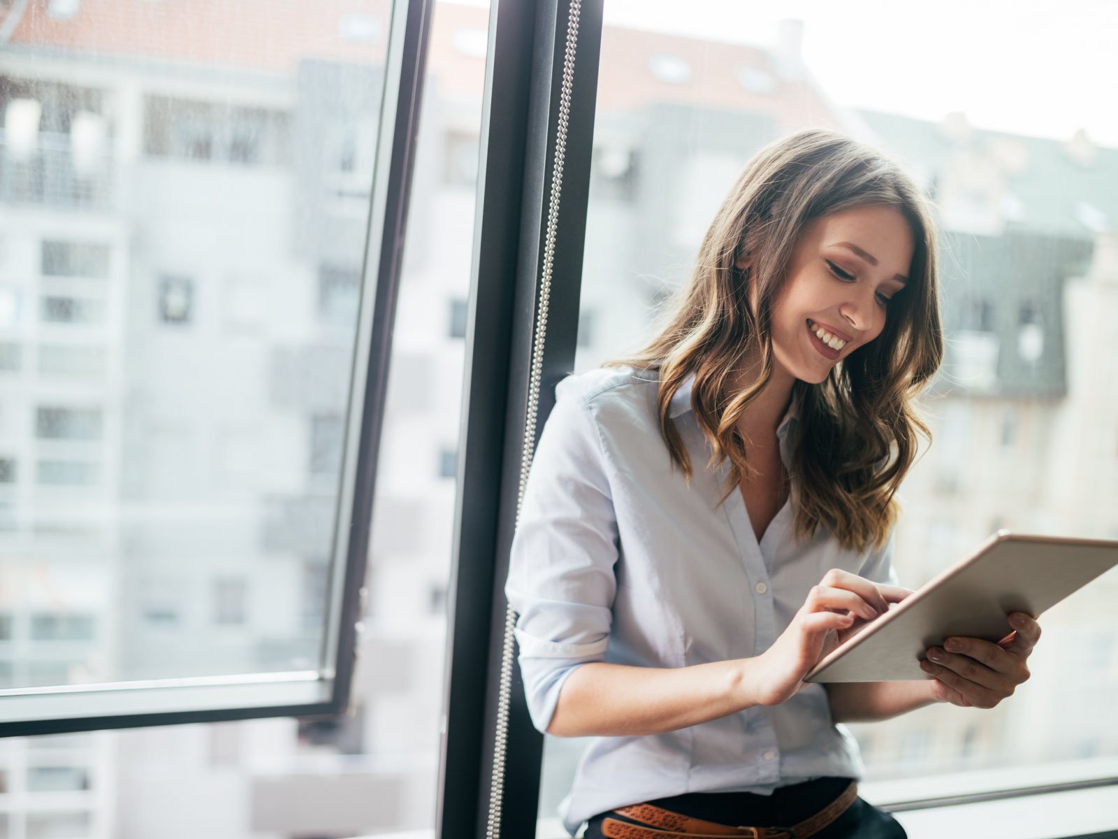 smiling woman in business setting looking at tablet leaning against window